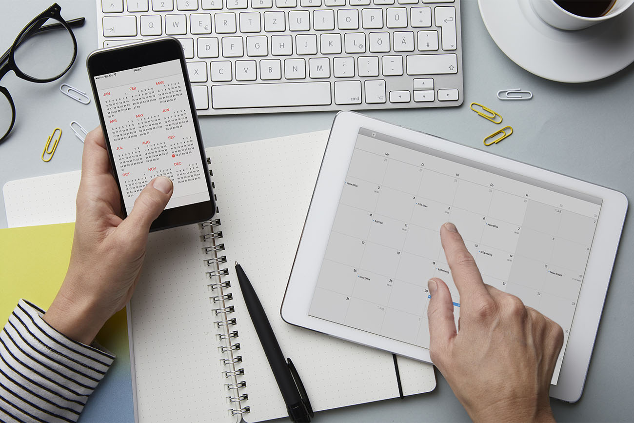 Aerial view of woman holding a smartphone and tablet both with calendars on an office desk | © Westend61 / Getty Images